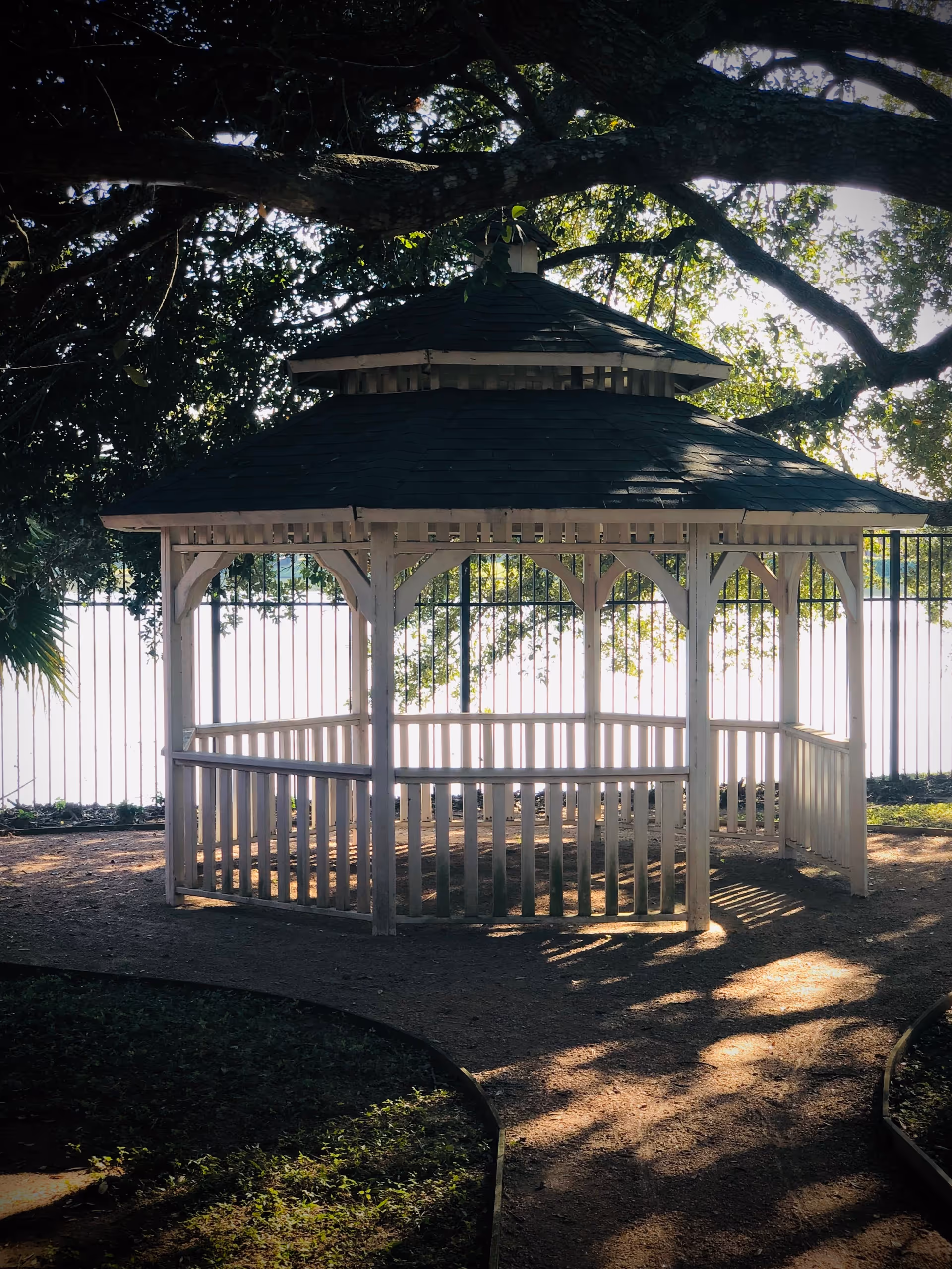 A white wooden gazebo with a dark shingled roof situated outdoors under large tree branches, with sunlight filtering through the leaves and a metal fence in the background.