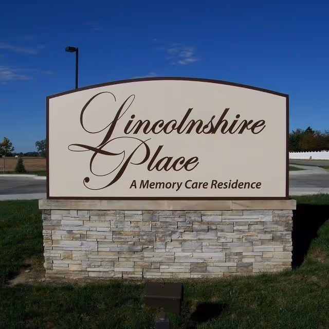 Large stone-based roadside sign reading "Lincolnshire Place — A Memory Care Residence" against a blue sky.