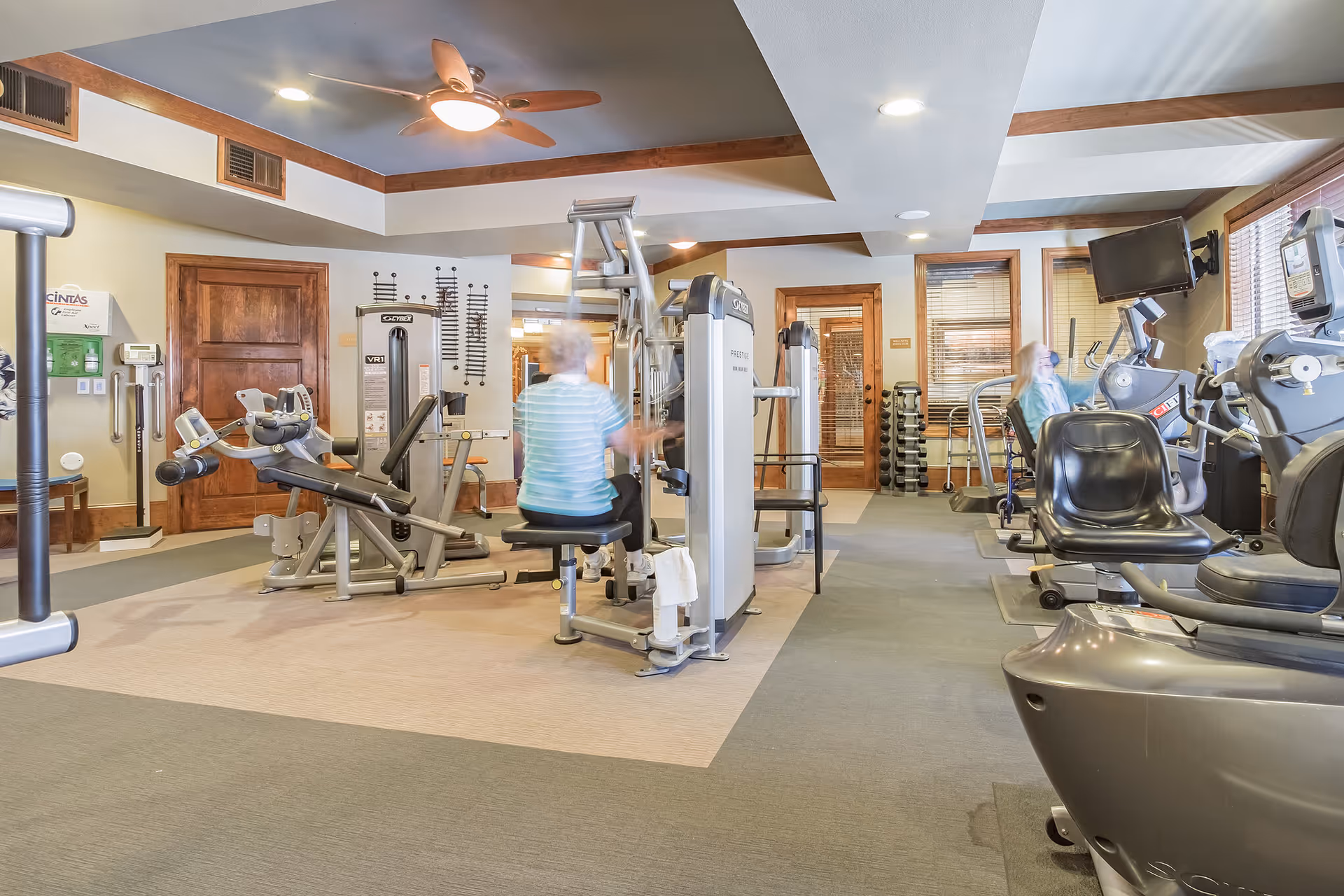 A fitness room in a senior living facility with various exercise machines including stationary bikes, weight machines, and treadmills. Two elderly individuals are using the equipment. The room has a ceiling fan, recessed lighting, and wooden trim around the doors and windows.