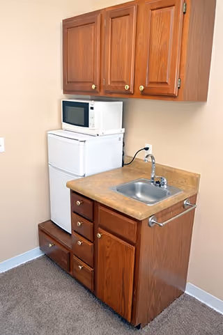 Small kitchenette area with wooden cabinets, a countertop with a sink, a white microwave on top of a white mini refrigerator, and a beige wall background.