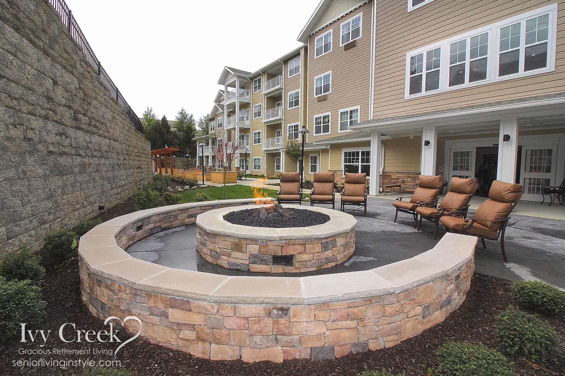 Outdoor courtyard with a circular stone fire pit, cushioned chairs, and a multi-story senior living building in the background.