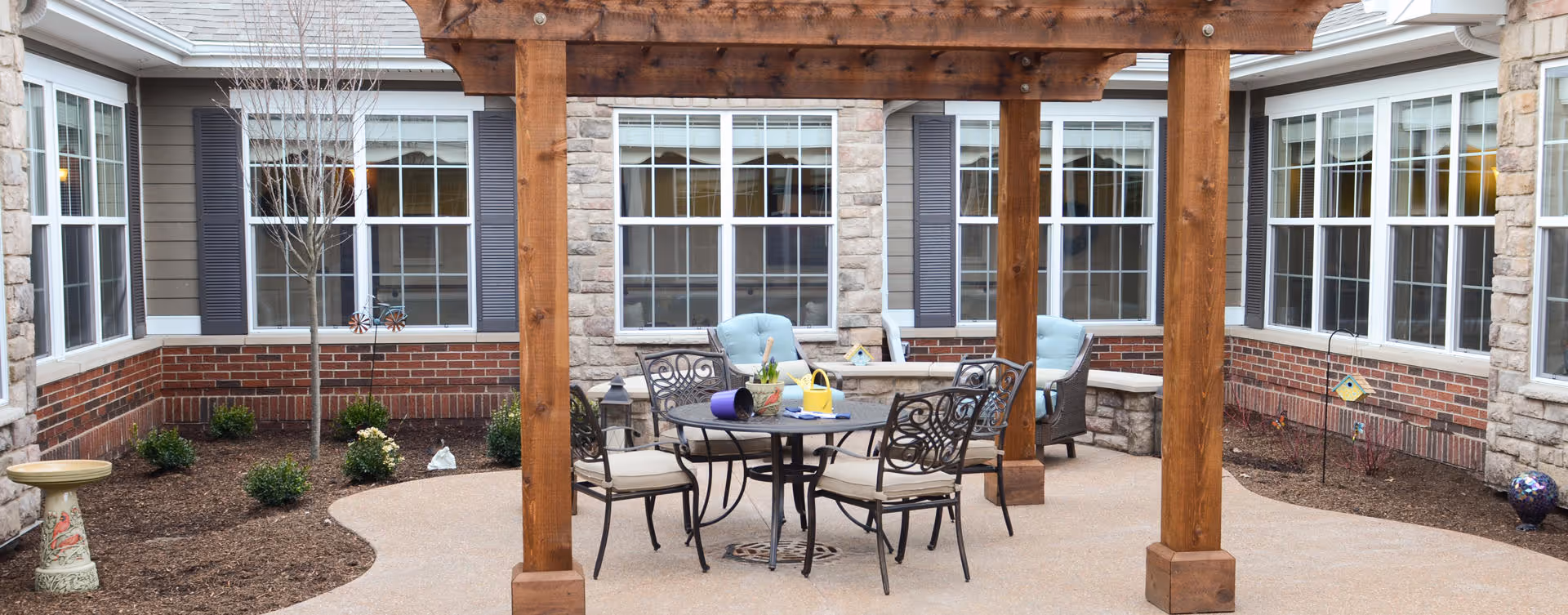 Outdoor patio area with a wooden pergola, a round metal table with four cushioned chairs, and two cushioned armchairs near a stone wall. The patio is surrounded by a garden bed with small shrubs and a birdbath, and the building has multiple large windows with white frames and dark shutters.