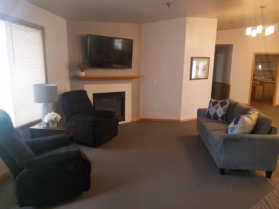 Cozy living room with two recliners, a sofa with patterned pillows, a wall-mounted TV above a fireplace, and a side table with a lamp and flowers.