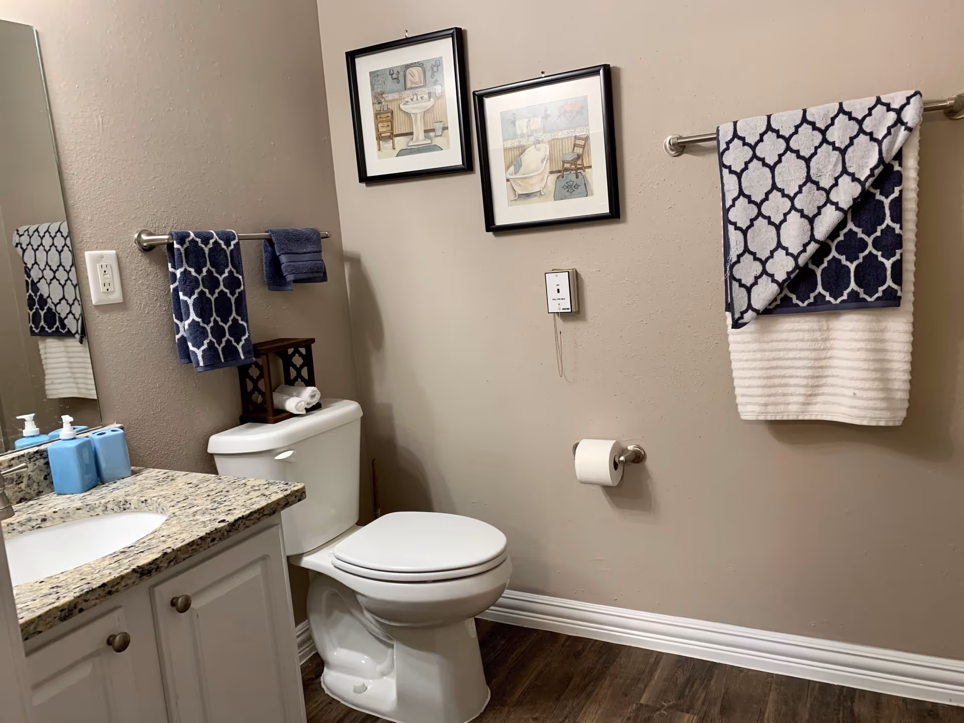 A clean bathroom with a white toilet, granite countertop with sink, blue soap dispensers, and patterned blue and white towels hanging on towel racks. Two framed pictures hang on the beige wall above the toilet, and a toilet paper holder is mounted on the wall. The floor has dark wood-style flooring.