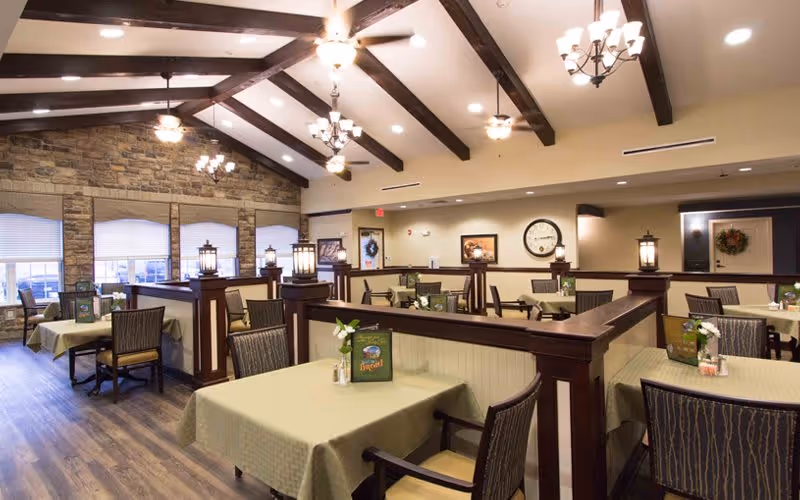 A spacious dining room with multiple tables covered in light green tablecloths, each set with chairs and small flower arrangements. The room features wooden beams on the ceiling, chandeliers, and large windows with blinds. The walls have stone accents and framed pictures, creating a warm and inviting atmosphere.