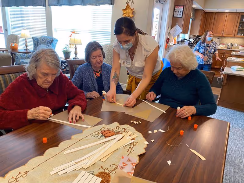 Three elderly women sitting at a wooden table engaged in a craft activity with paper strips and glue sticks, assisted by a caregiver wearing a mask. Another staff member is visible in the background near a kitchen area. The room is well-lit with windows and decorated with homey touches.