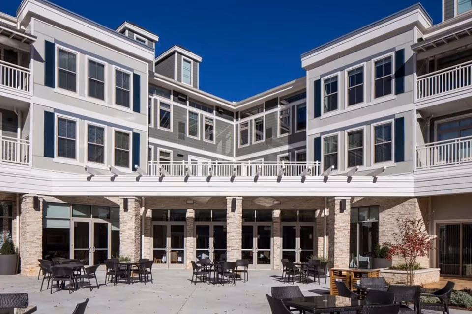 Outdoor courtyard area of a senior living facility with multiple black tables and chairs arranged on a concrete patio. The building surrounding the courtyard is three stories tall with large windows, white and gray exterior walls, and balconies. The sky is clear and blue.