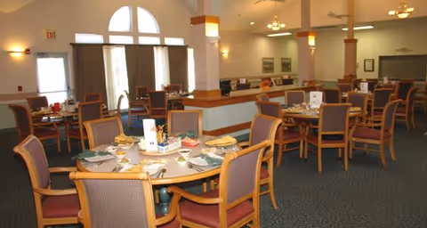 Spacious dining room with multiple round tables set with plates, napkins, and utensils. Chairs with wooden frames and cushioned seats surround the tables. Large windows with curtains allow natural light into the room. The room has high ceilings with ceiling fans and light fixtures, and a carpeted floor.