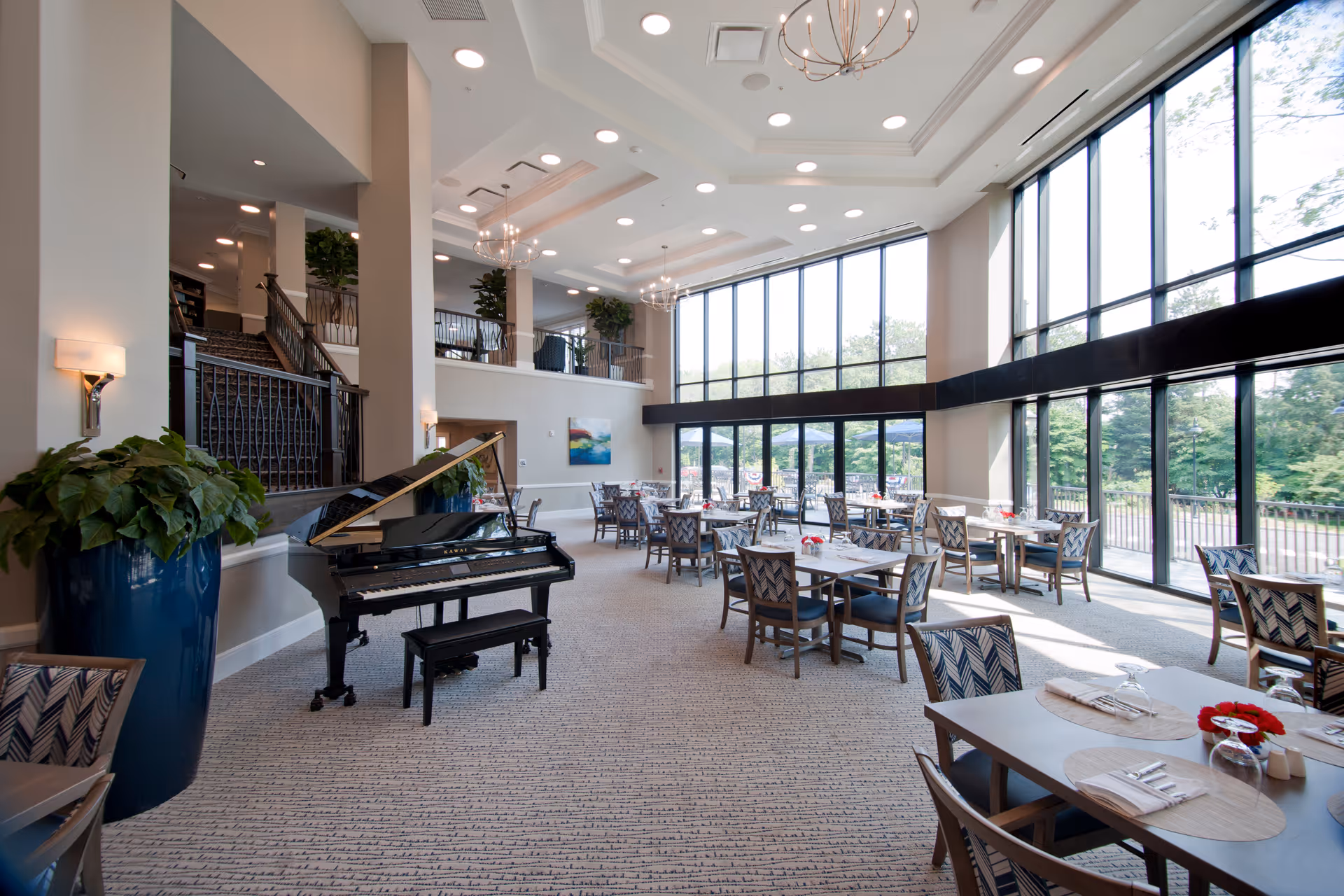 A spacious and bright dining area with large floor-to-ceiling windows allowing natural light to fill the room. The room features multiple tables with chairs arranged neatly, each table set with placemats, napkins, and glassware. A black grand piano with a matching bench is positioned near a large blue planter with green plants. The space has high ceilings with modern light fixtures and a staircase leading to an upper level with additional seating and plants.