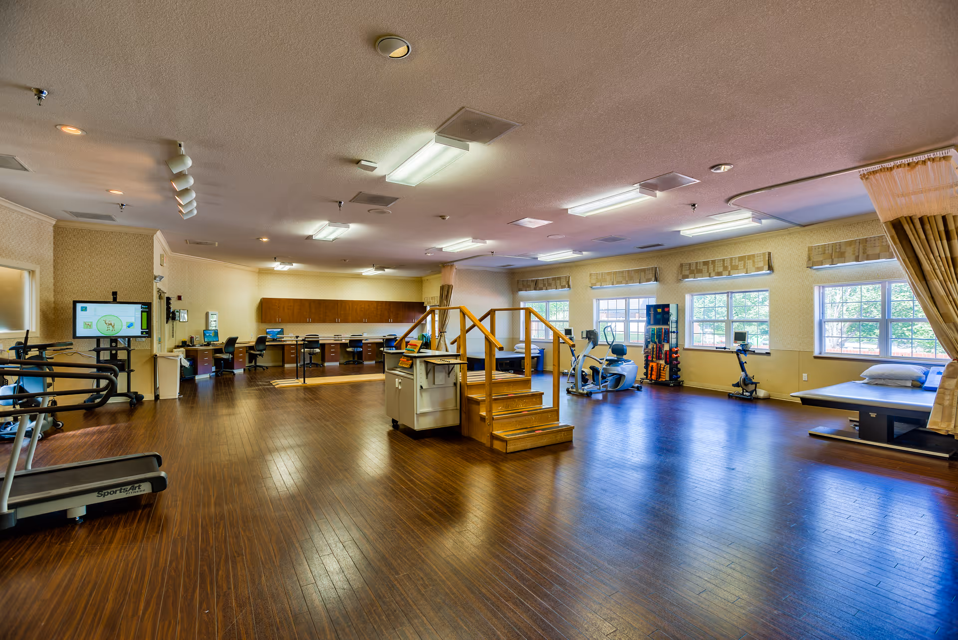 A spacious rehabilitation room with wooden flooring, exercise equipment including a treadmill, stationary bikes, and a set of stairs with handrails for physical therapy. There are several desks and chairs along the back wall, large windows letting in natural light, and a curtained area with a therapy bed.