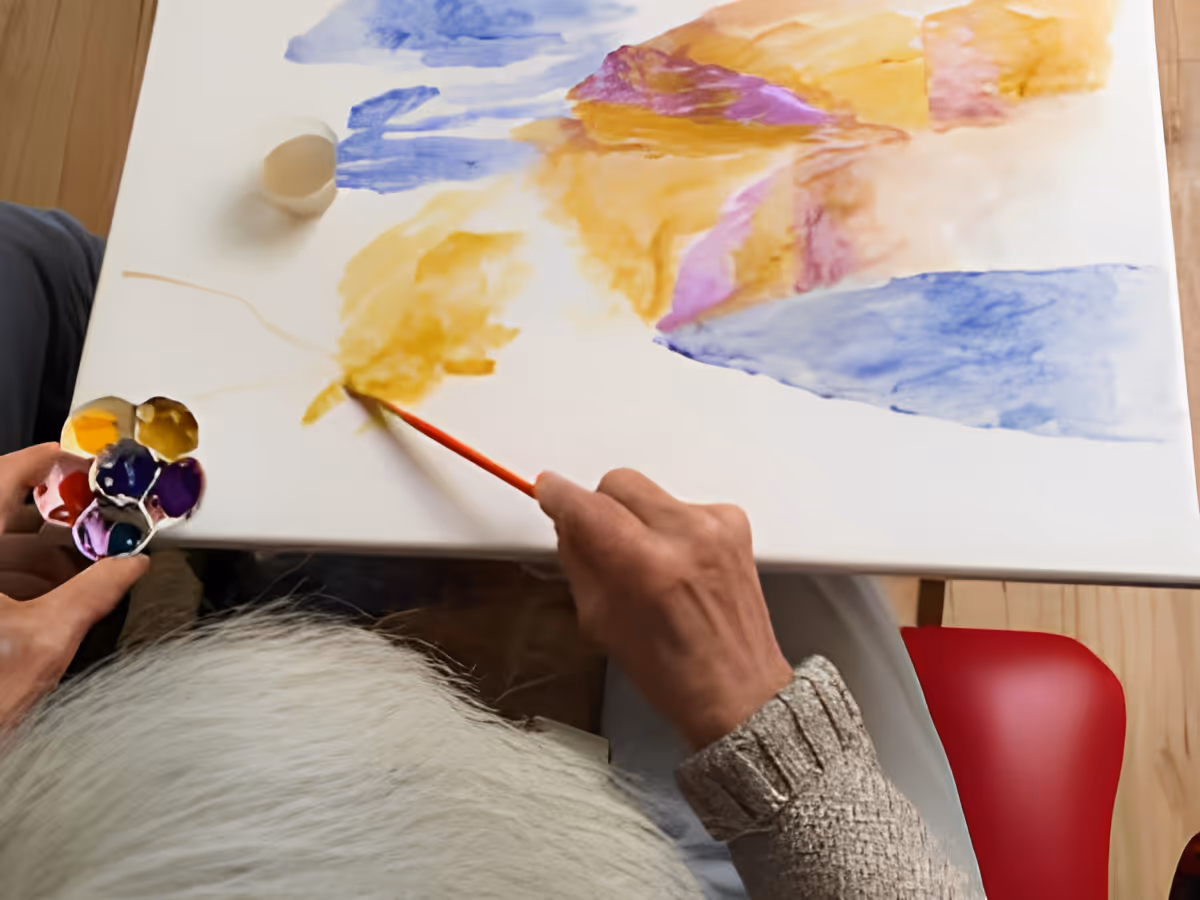 Overhead view of a person painting a colorful abstract watercolor artwork on a white canvas, holding a small palette with various paint colors in one hand and a paintbrush in the other.