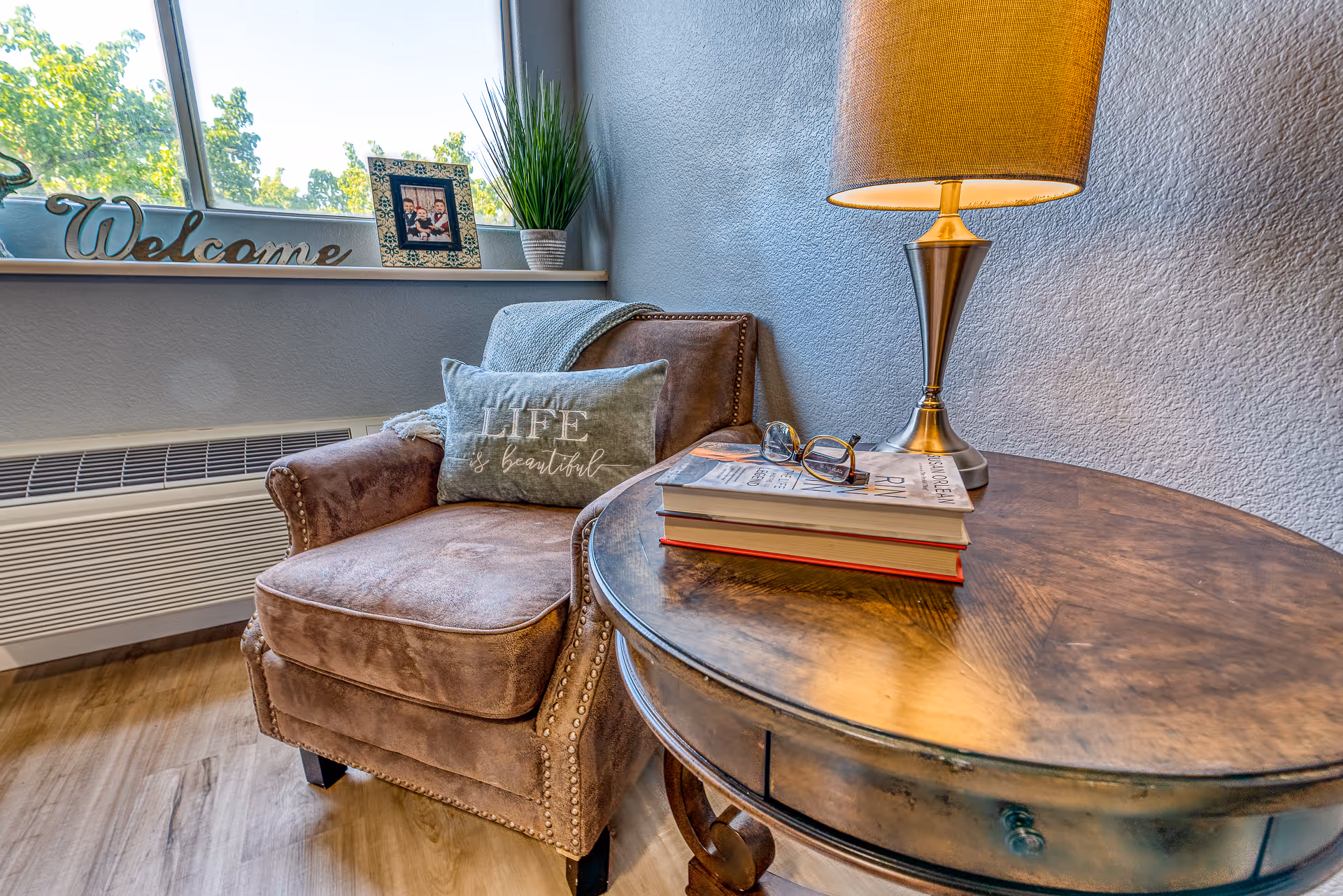 Cozy corner with a brown upholstered armchair featuring a decorative pillow that says 'LIFE is beautiful'. Next to the chair is a round wooden side table with a stack of books, a pair of glasses, and a lit table lamp. A window sill behind the chair holds a 'Welcome' sign, a framed photo, and a potted plant. The room has light-colored wooden flooring and textured light gray walls.