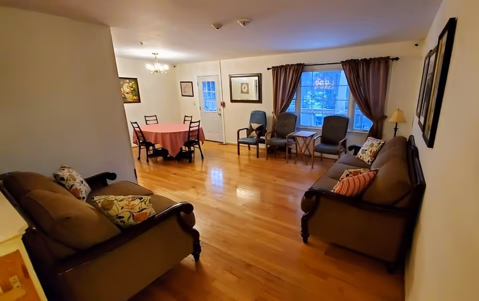 Spacious communal living room with sofas, chairs, a small dining table, hardwood floors and a large window with curtains.