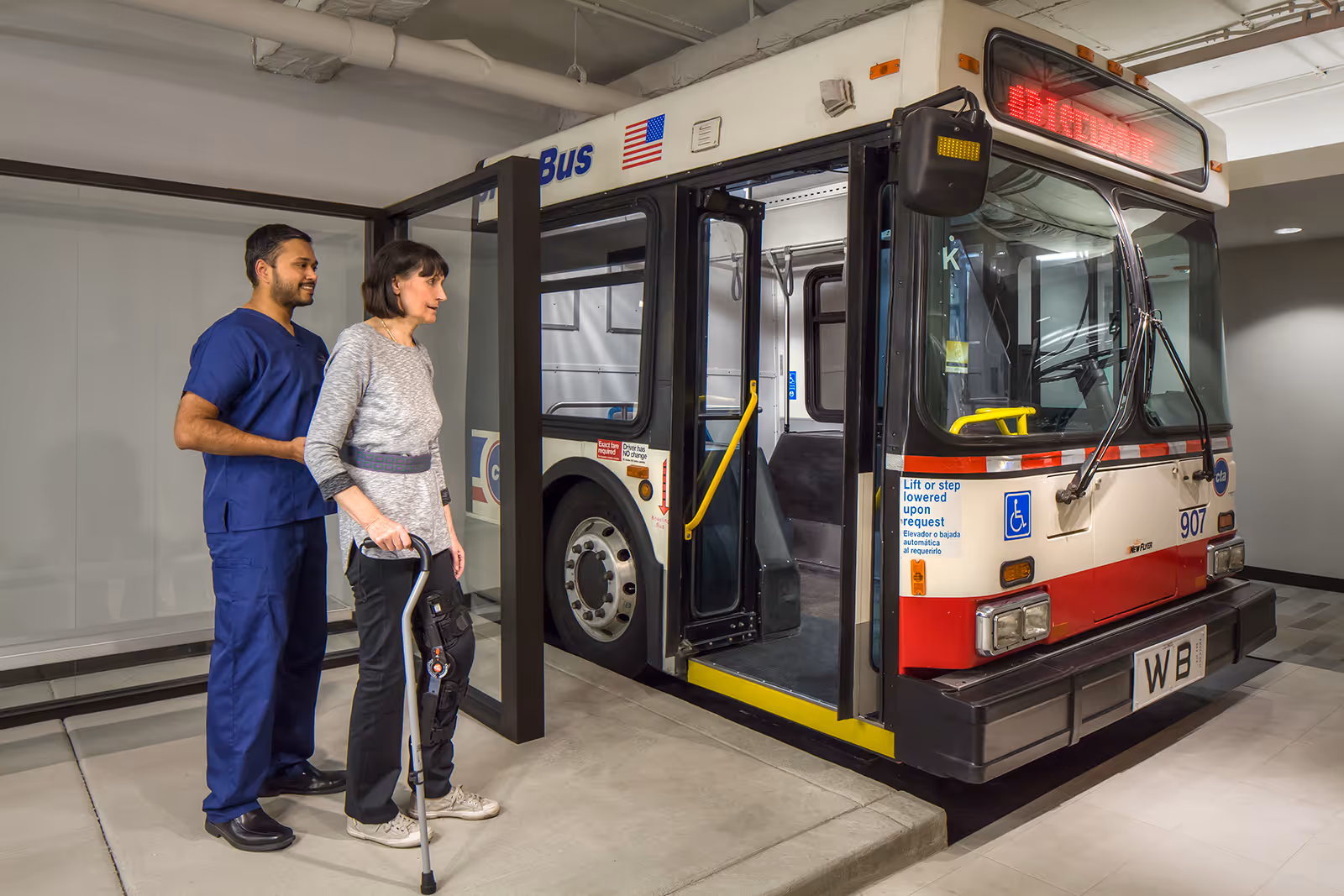 A healthcare worker in blue scrubs assists an elderly woman with a knee brace and cane as she prepares to board a stationary bus inside a building.