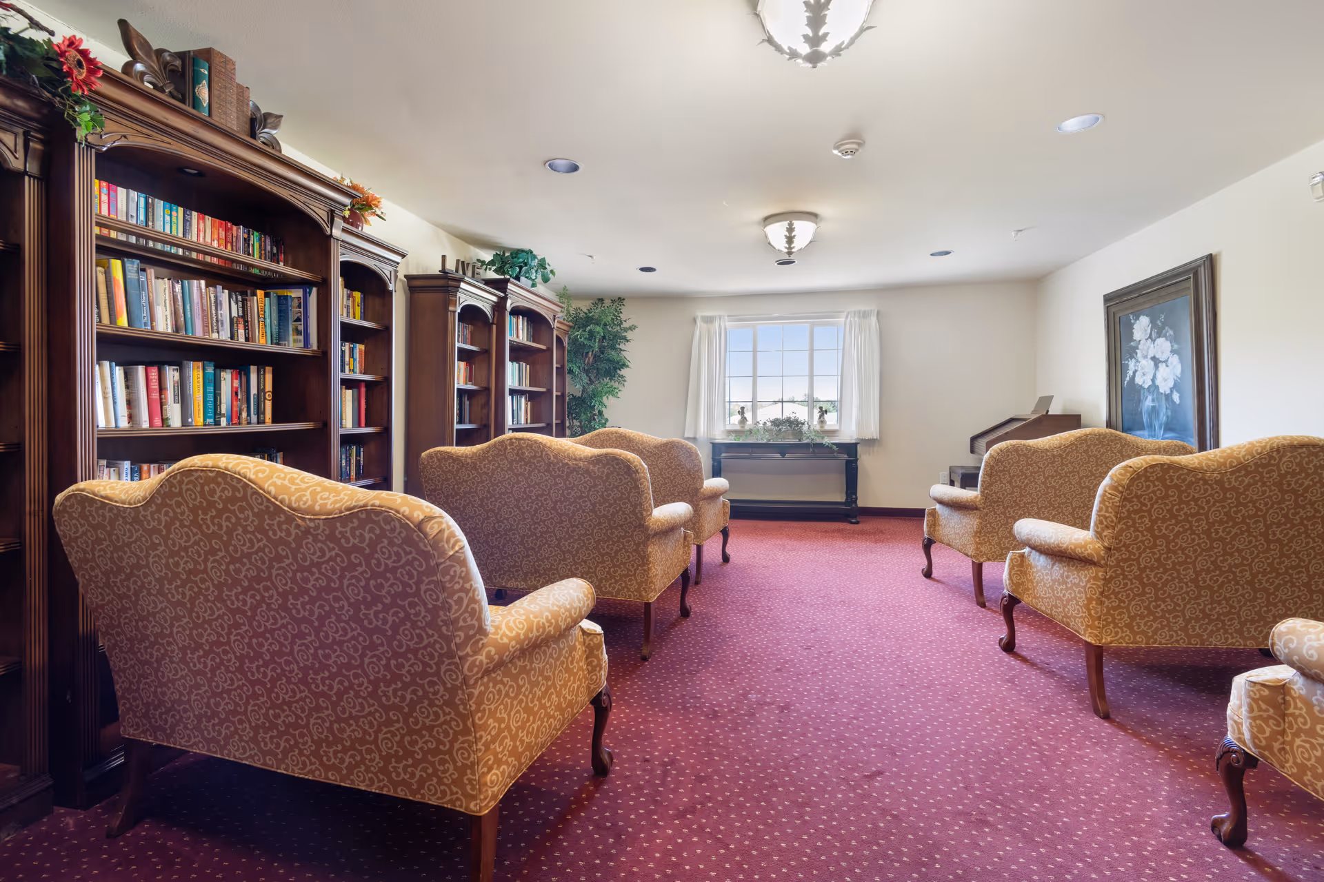 A cozy senior living community library or lounge area with several upholstered armchairs arranged in two rows facing a window. The room features wooden bookshelves filled with books along the left wall, a red carpeted floor, a window with white curtains, a framed floral painting on the right wall, and ceiling lights.