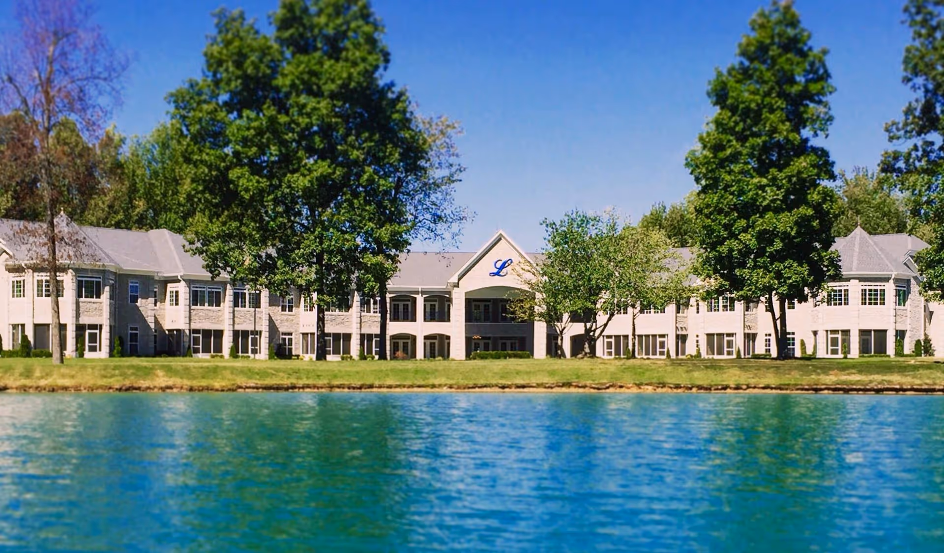 Two-story senior living building with trees along its lawn set beside a blue lake under a clear sky.