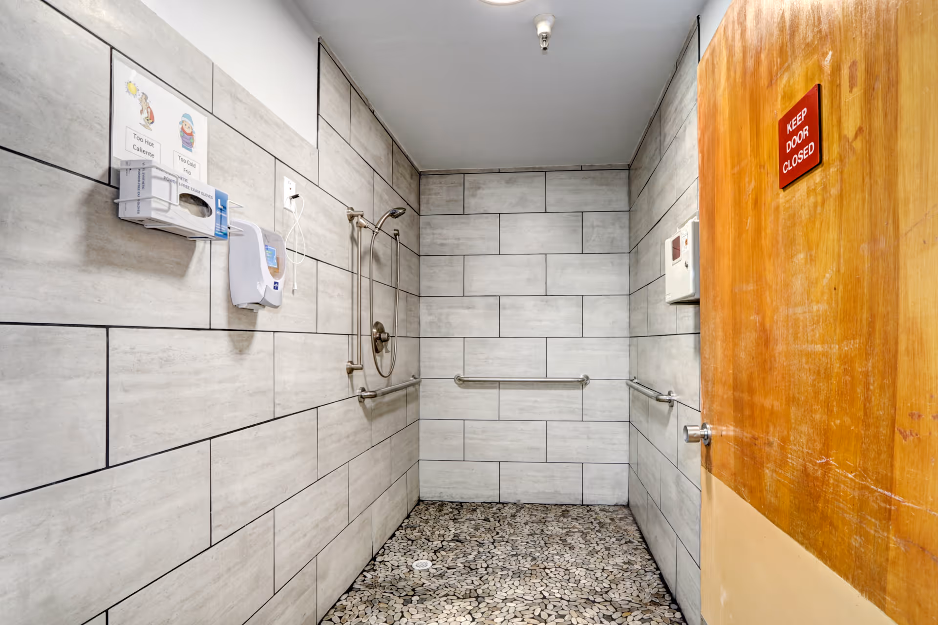 Accessible tiled shower room with grab bars, a handheld showerhead, soap dispensers and a wooden door with a 'KEEP DOOR CLOSED' sign.