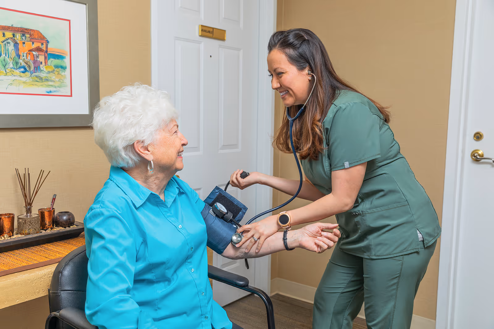 A healthcare worker in green scrubs is smiling while taking the blood pressure of an elderly woman with white hair who is seated and wearing a turquoise shirt. They are in a room with beige walls, a door, and a framed painting on the wall.