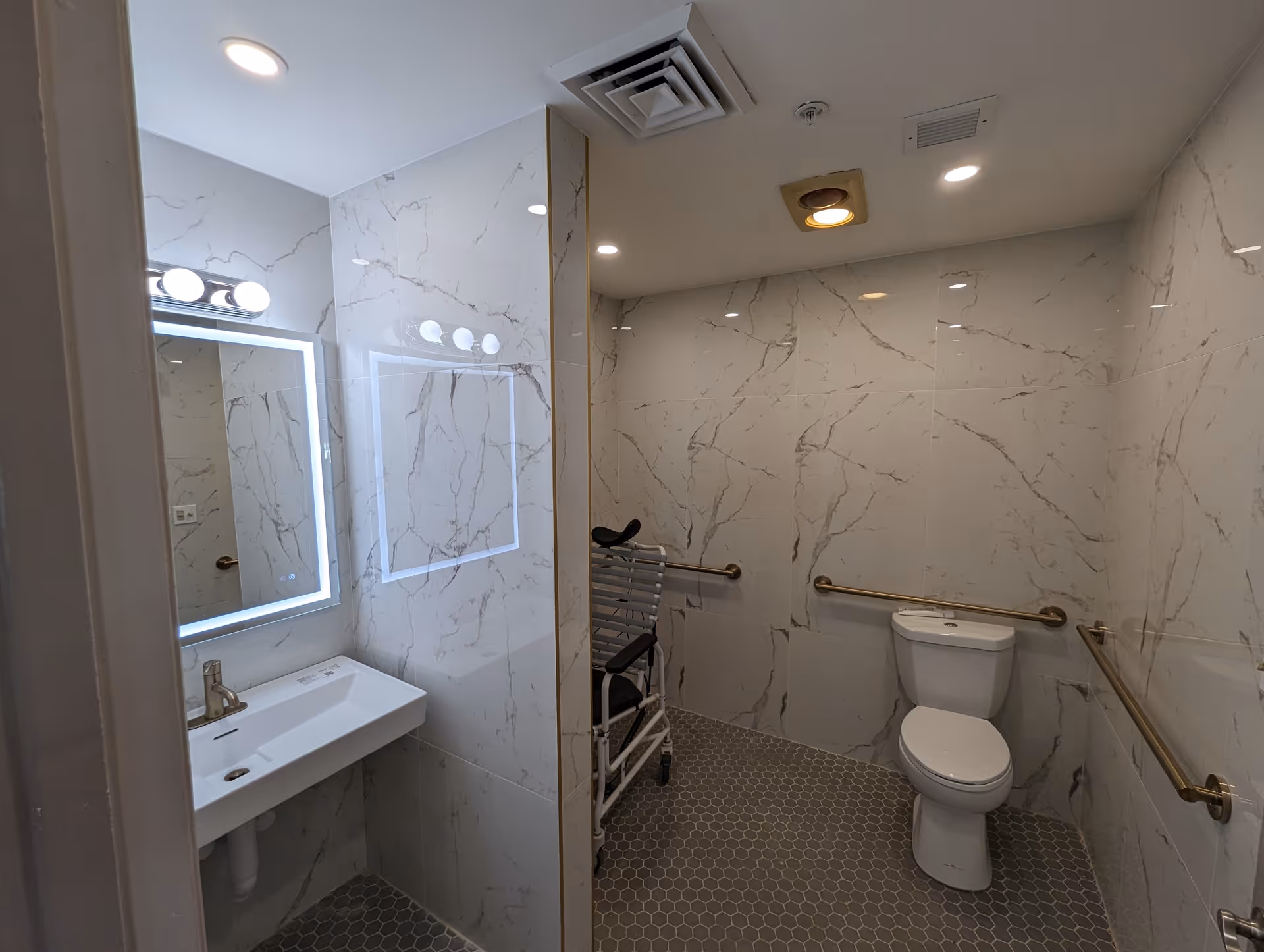 A modern, accessible bathroom with marble-patterned walls and hexagonal floor tiles. The bathroom features a white toilet with grab bars on both sides, a wall-mounted sink with a lit mirror above it, and a shower chair positioned near the toilet. The ceiling has recessed lighting and ventilation.