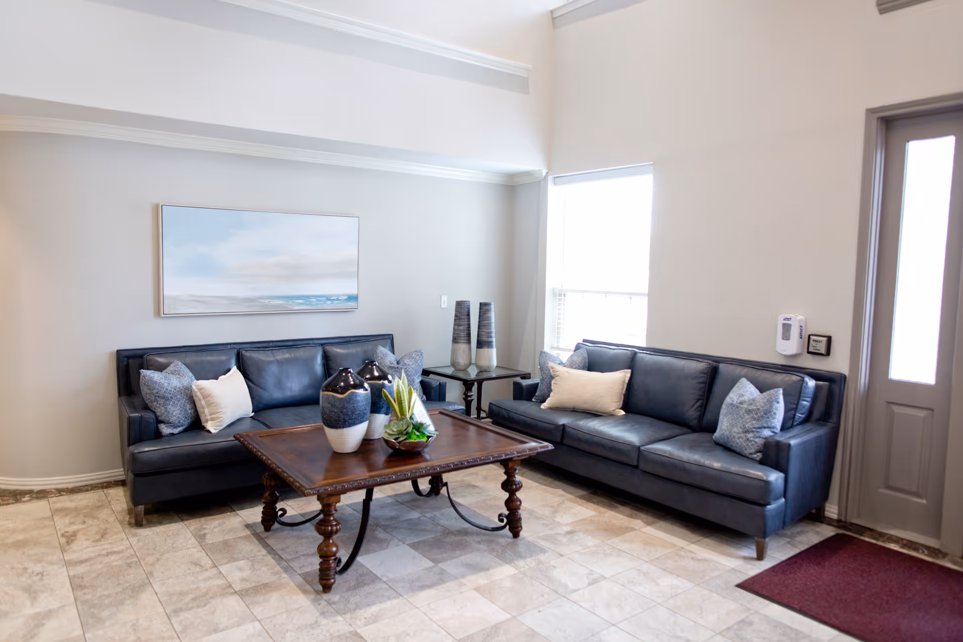 A bright seating area with two dark leather sofas, a wooden coffee table with decorative vases, and a window near the entrance.