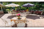 Outdoor patio area with several metal tables and chairs arranged on a tiled floor. Two large umbrellas provide shade over some tables. There are potted plants with flowers on the tables and greenery in the background.