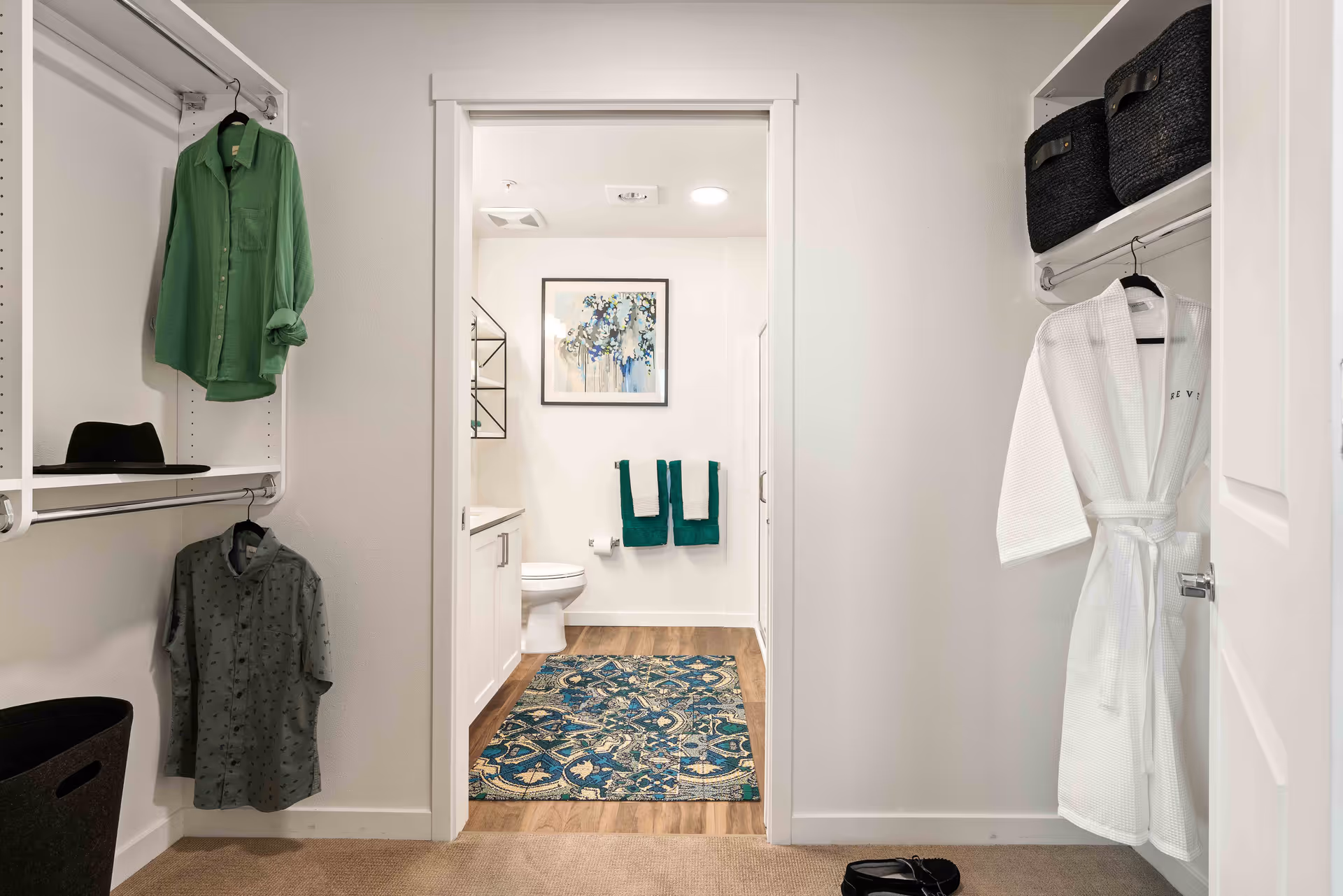 View from a walk-in closet into a bathroom. The closet has hanging clothes including a green shirt, a gray shirt, and a white robe. There are black storage baskets on shelves and a black hat on a shelf. The bathroom has a toilet, a vanity with a sink, two green and white towels hanging on the wall, a framed abstract artwork, and a blue patterned rug on the wooden floor.