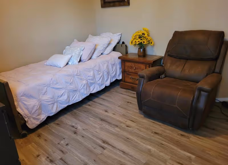 A small bedroom with a single bed covered in a light purple quilt and several pillows. Next to the bed is a wooden nightstand with a vase of yellow flowers. To the right of the nightstand is a large brown recliner chair. The floor is wood laminate and the walls are light beige.