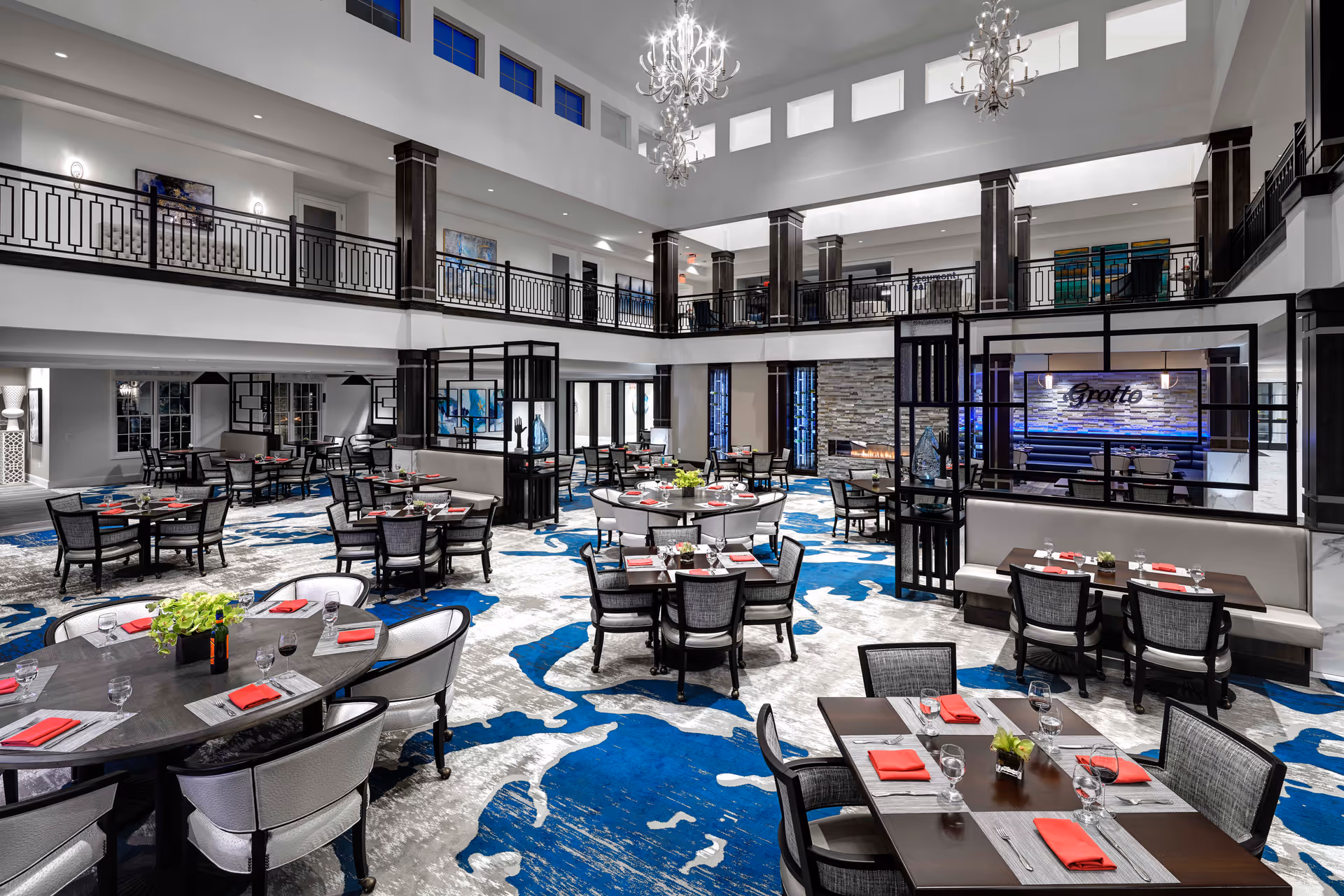 Spacious and modern dining room with multiple round and rectangular tables set with glasses, plates, and red napkins. The room features a high ceiling with chandeliers, a patterned blue and gray carpet, and a second-floor balcony overlooking the area. There is a fireplace and a sign that reads 'Grotto' on one wall.