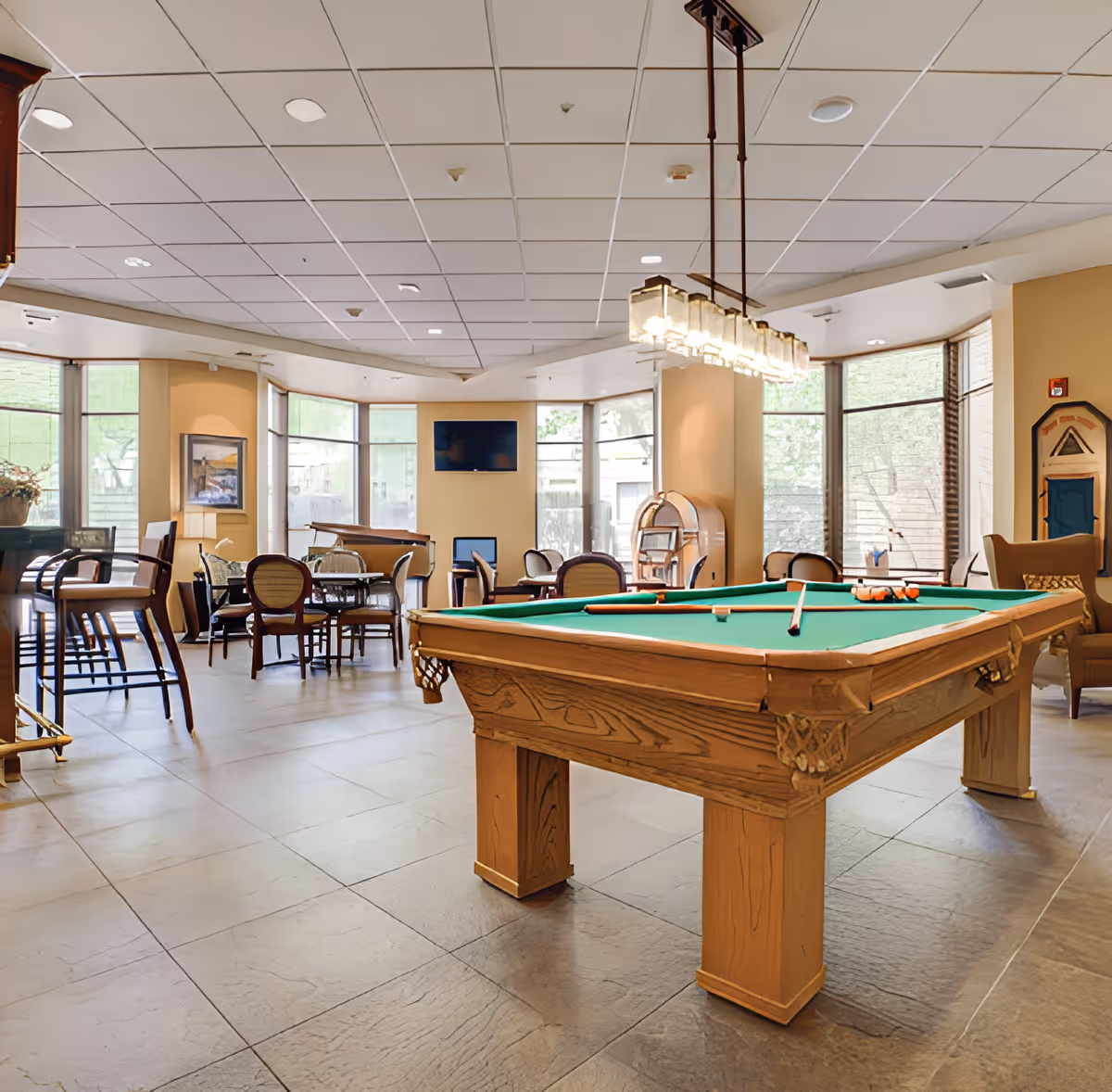 A spacious recreational room with a pool table in the foreground, surrounded by chairs and tables. Large windows allow natural light to fill the room. A hanging light fixture is above the pool table, and a jukebox is visible near the back wall.
