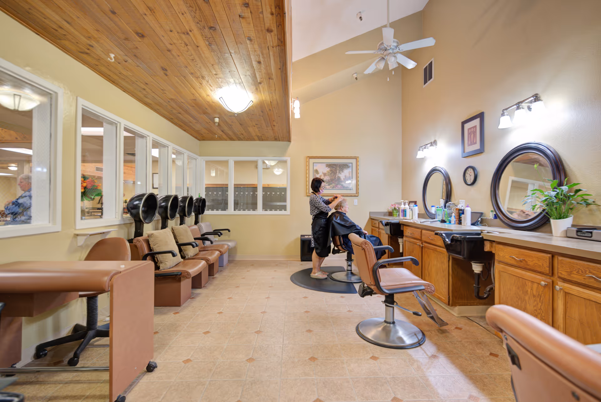 Interior view of a hair salon area in a retirement community with a stylist cutting an elderly woman's hair. The room features wooden cabinets with mirrors and various hair products on the counter. There are multiple salon chairs and hair drying stations along the left wall. The ceiling has wooden paneling and a ceiling fan.