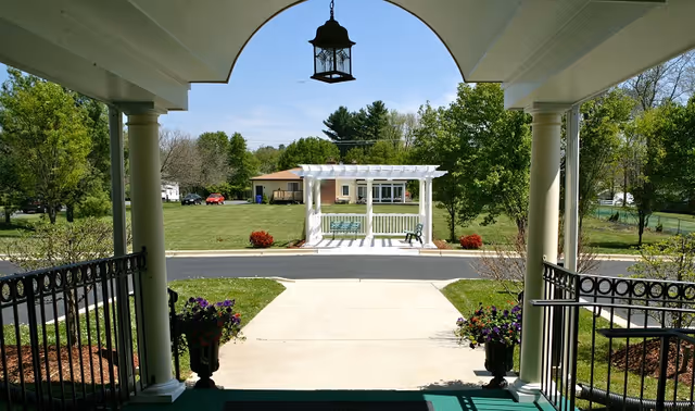 View from a covered entrance with columns and a hanging lantern looking out over a paved walkway to a white pergola on a grassy lawn.