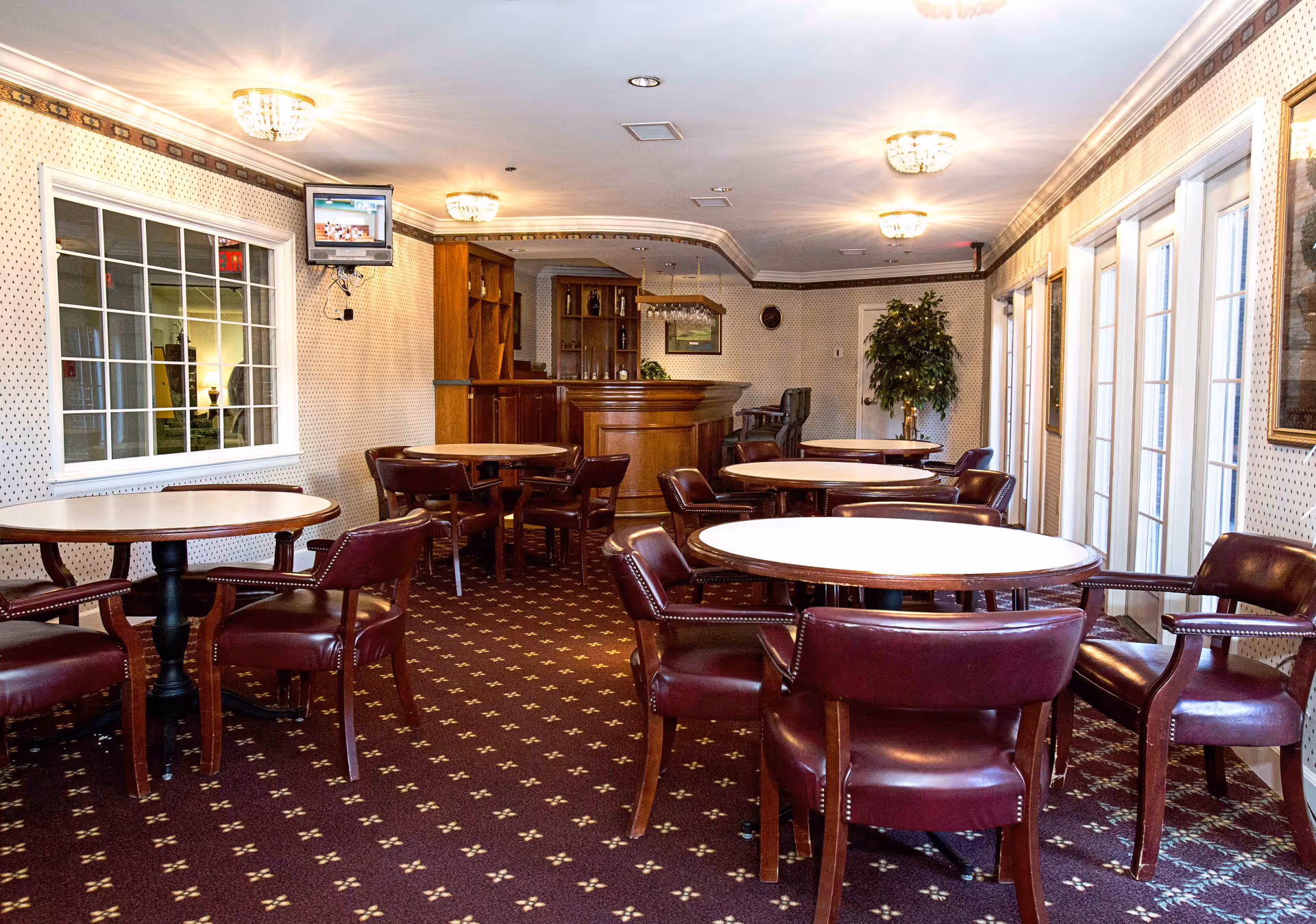 A cozy dining room with several round tables surrounded by maroon leather chairs. The room has patterned carpet and wallpaper, a small wooden bar area in the back, a wall-mounted TV, and large windows with white frames letting in natural light.
