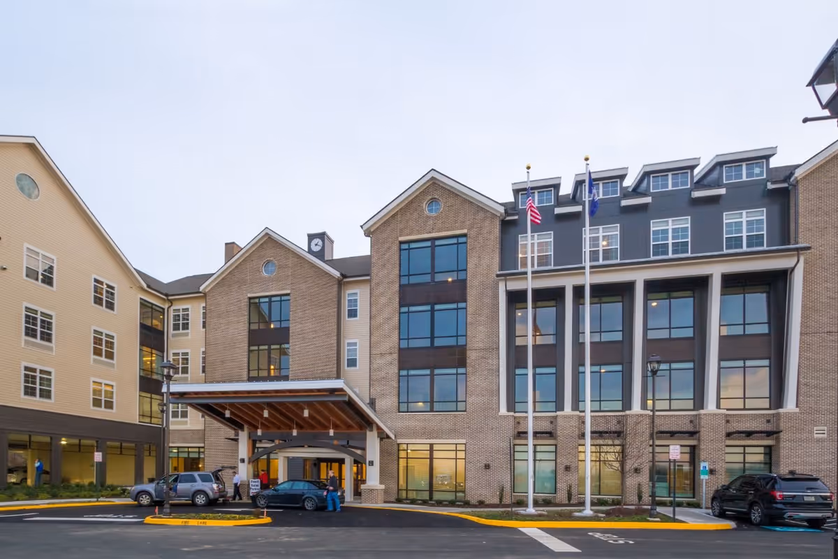 Front exterior view of Tribute at The Glen senior living facility showing a multi-story building with large windows, a covered entrance, two flagpoles with flags, and a parking area with several cars.
