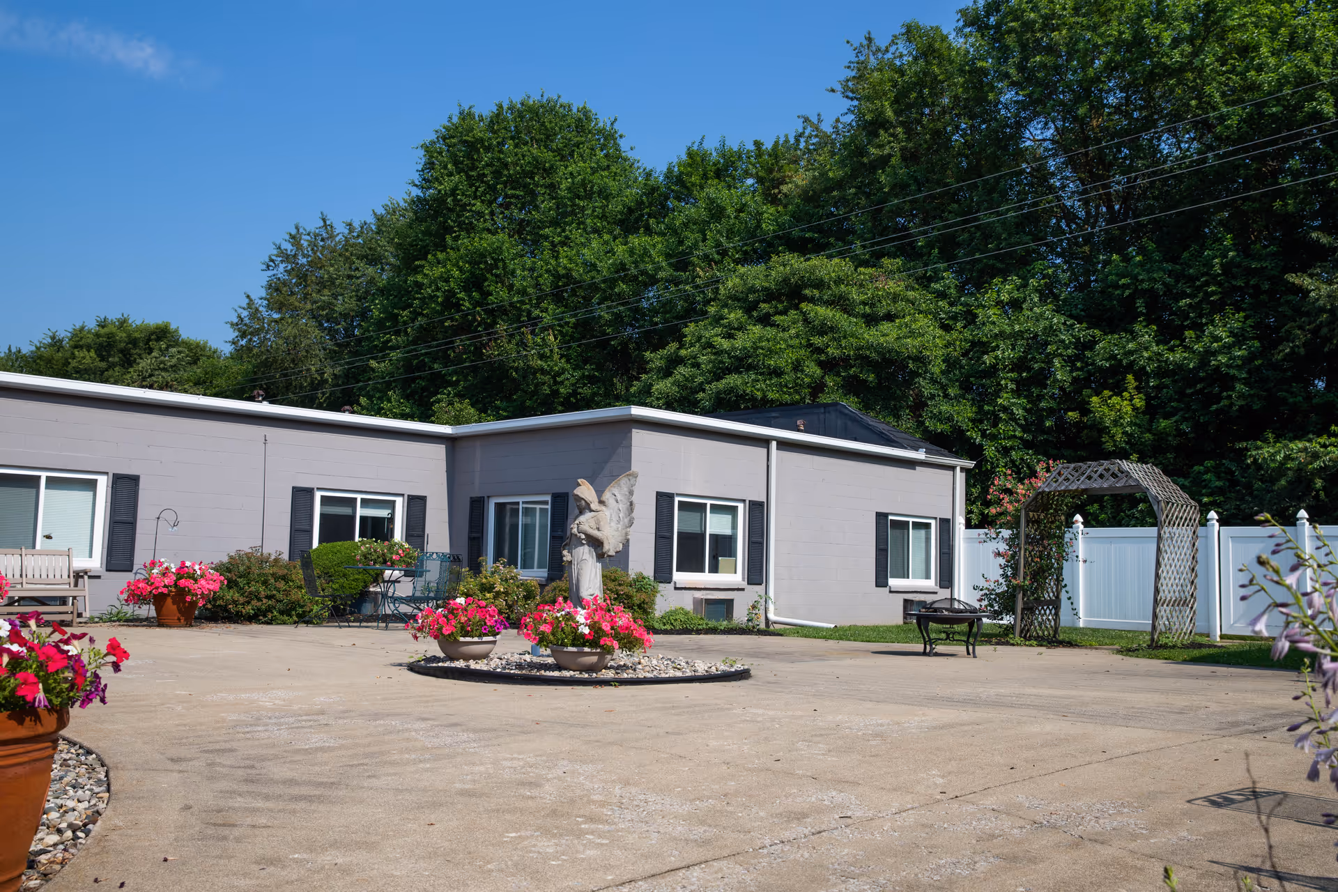 Outdoor courtyard area of a nursing and rehabilitation facility with a concrete ground, flower pots with pink flowers, a statue of an angel, a wooden bench, a small table with chairs, a white fence, and green trees in the background under a clear blue sky.