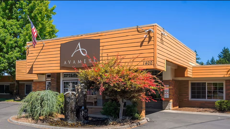 Exterior front view of Avamere Rehabilitation of Oregon City building with a large sign displaying the facility name, an American flag, trees, shrubs, and a small water fountain in front under a clear blue sky.