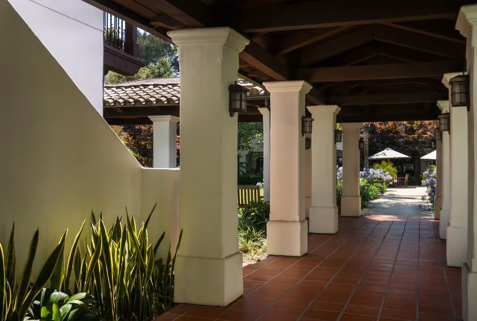 Covered outdoor walkway with white columns and terracotta tiled floor, surrounded by greenery and leading to a garden area with benches and umbrellas.