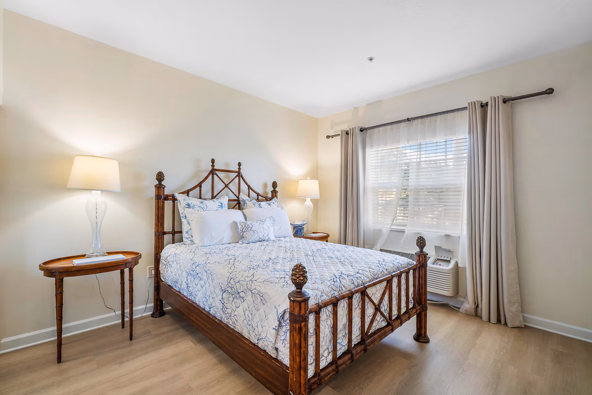 A bright bedroom with a wooden bed frame featuring a white and blue patterned bedspread and multiple pillows. There are two bedside tables, each with a lamp. A window with sheer white curtains and beige drapes lets in natural light, and the floor is light wood.