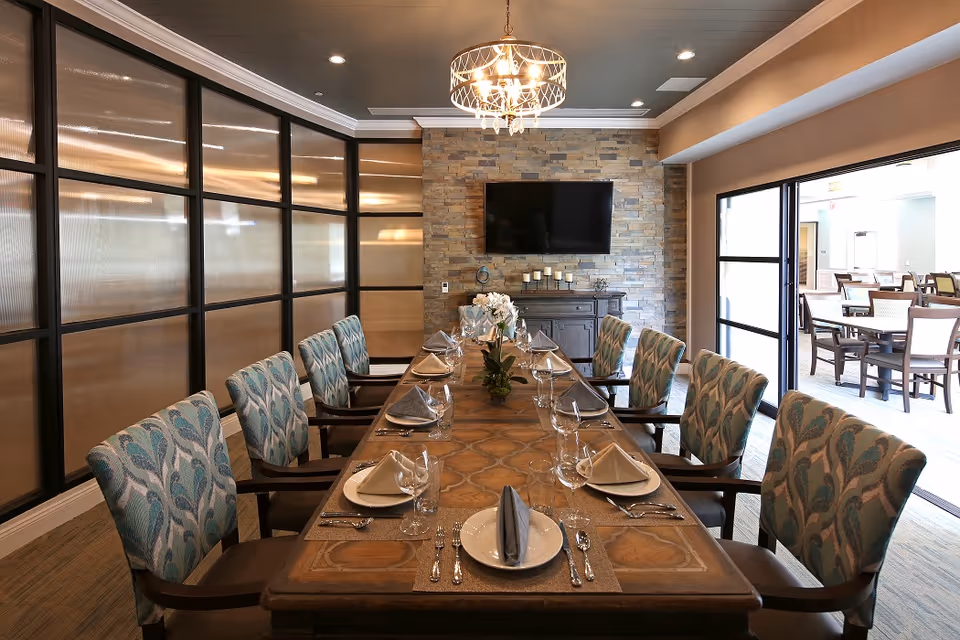 A long wooden dining table set for a meal with plates, napkins, glasses, and silverware. The table is surrounded by upholstered chairs with a blue and green pattern. A chandelier hangs above the table, and a flat-screen TV is mounted on a stone accent wall at the far end. Large windows and glass doors allow natural light into the room, revealing another dining area beyond.