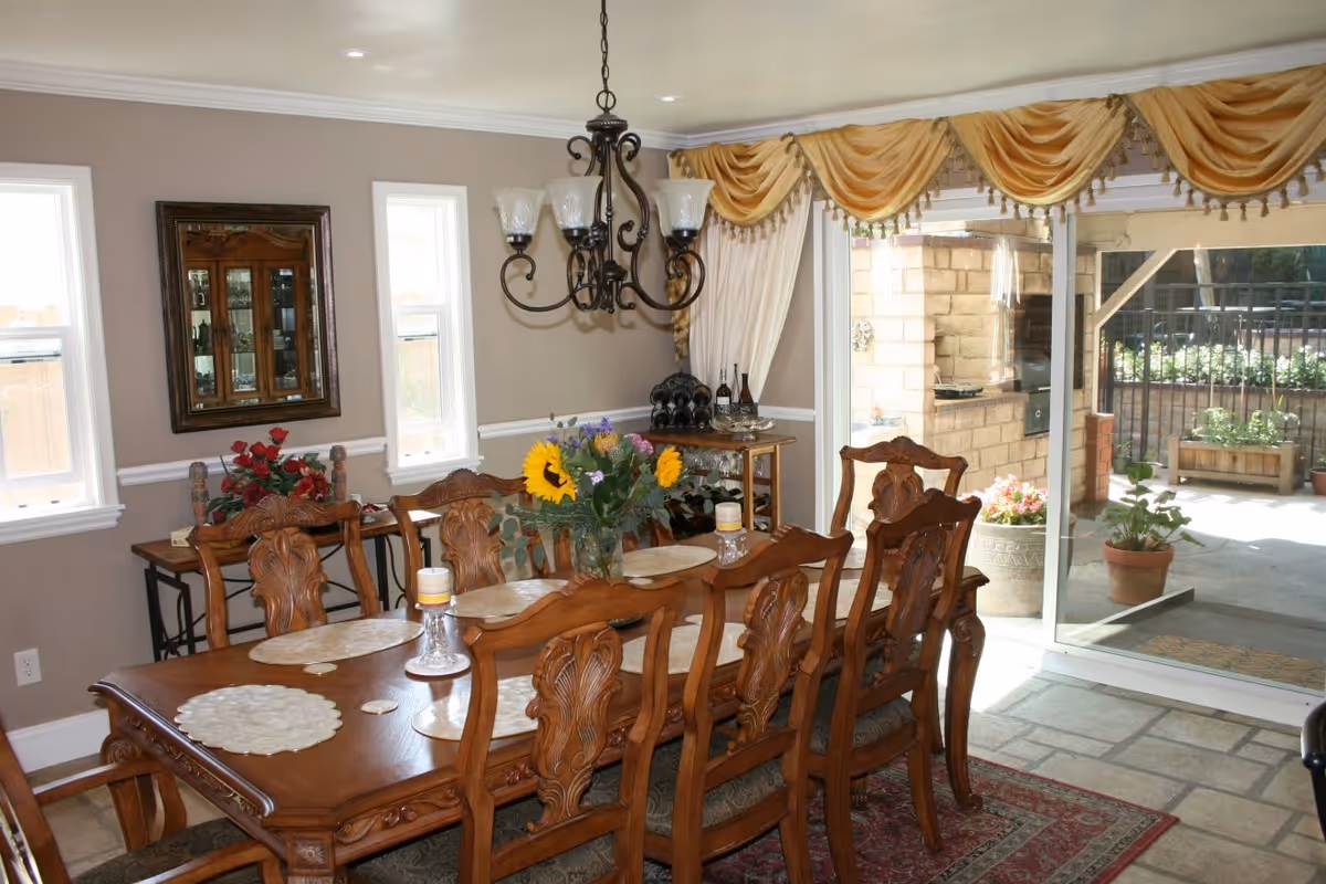 A dining room with a large wooden dining table surrounded by eight ornate wooden chairs. The table is decorated with placemats, candles, and a vase of flowers including sunflowers. There is a chandelier hanging above the table. The room has beige walls with white trim, two narrow windows, a mirror on the wall, and a side table with wine bottles. Large sliding glass doors with gold drapes open to an outdoor patio area with potted plants and a stone floor.