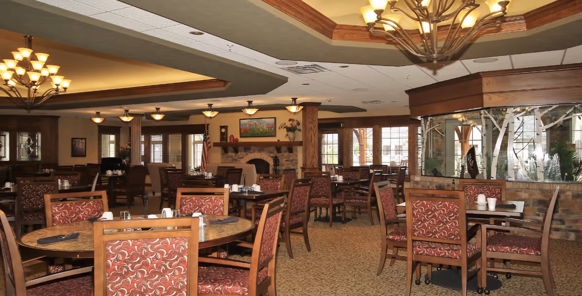 Spacious dining room with round wooden tables and upholstered chairs, chandeliers, and a fireplace in a senior living facility.