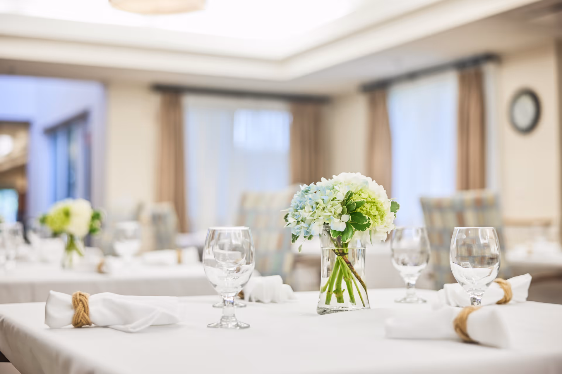 A dining table set with white tablecloth, neatly folded white napkins tied with twine, clear water glasses, and a small glass vase with a bouquet of white and green flowers in a softly lit dining room with blurred background.