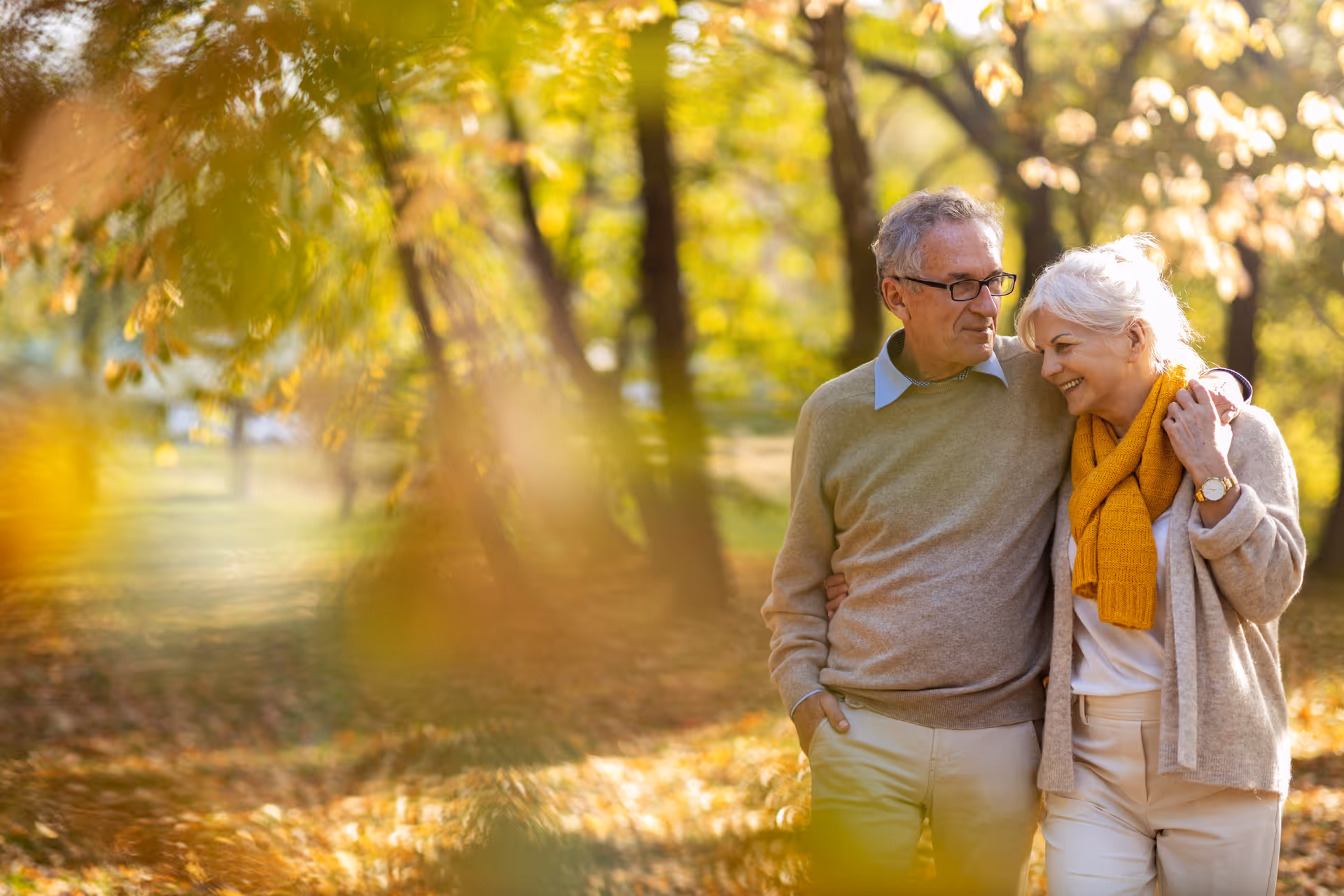 An older couple walking arm-in-arm through a sunlit park with autumn leaves and trees.