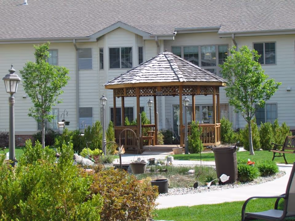 Outdoor garden area at Hillcrest Silver Ridge featuring a wooden gazebo with a shingled roof, surrounded by green trees, bushes, and a paved walkway. There are benches and lamp posts around the garden, with a multi-story building in the background.