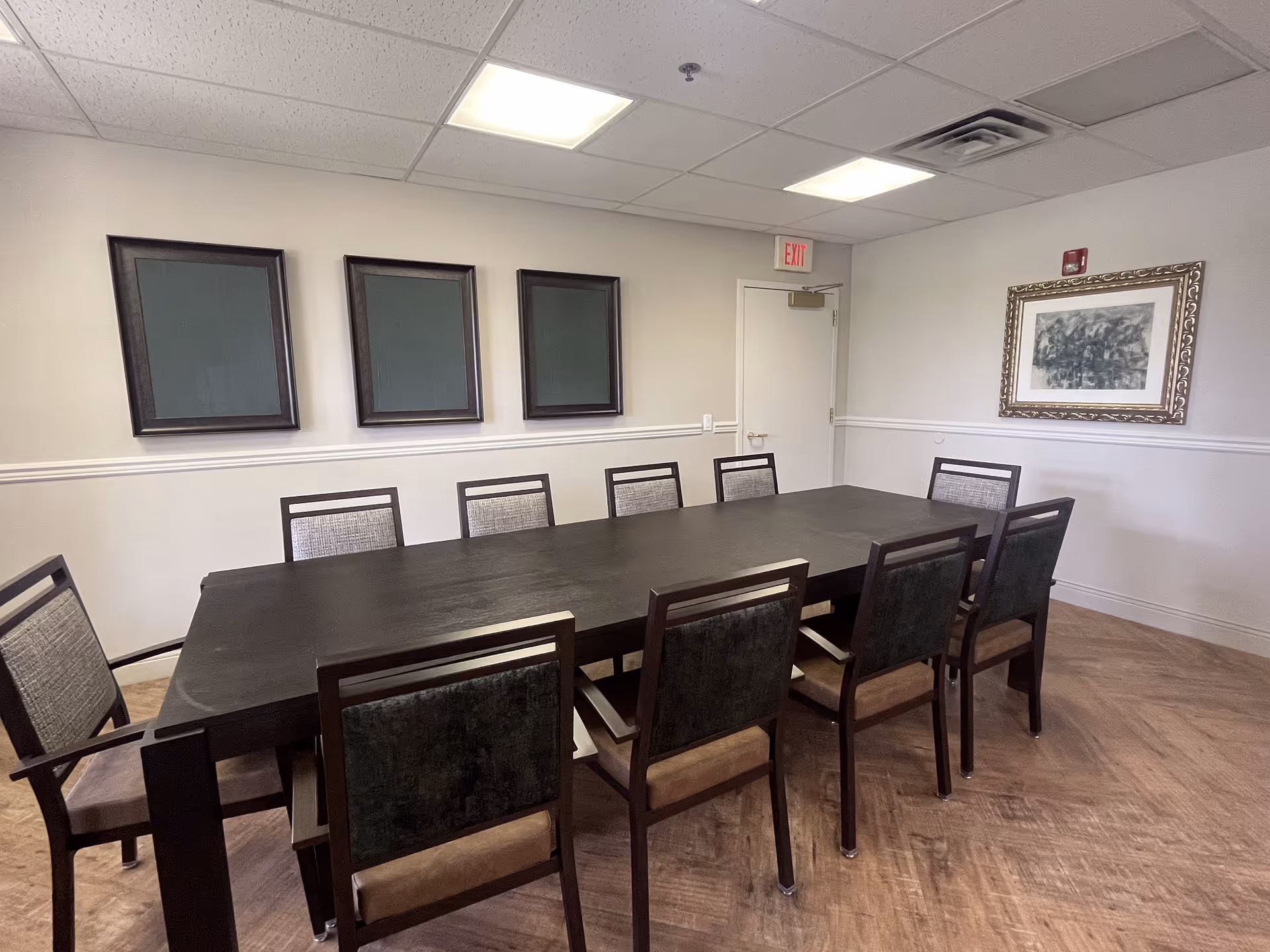 A conference or meeting room with a long dark wooden table surrounded by ten chairs. The walls are light-colored with three empty picture frames on one wall and a framed artwork on another. The ceiling has recessed lighting and a visible exit door is at the back of the room.