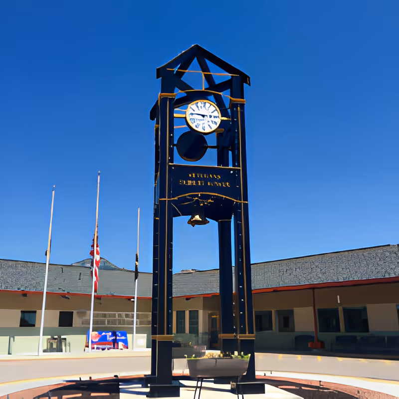 A tall black clock tower with a bell underneath the clock face stands in an outdoor courtyard area of the George E. Wahlen Ogden Veterans Home. The sky is clear and blue, and there are flagpoles with flags in the background near a single-story building with a flat roof.