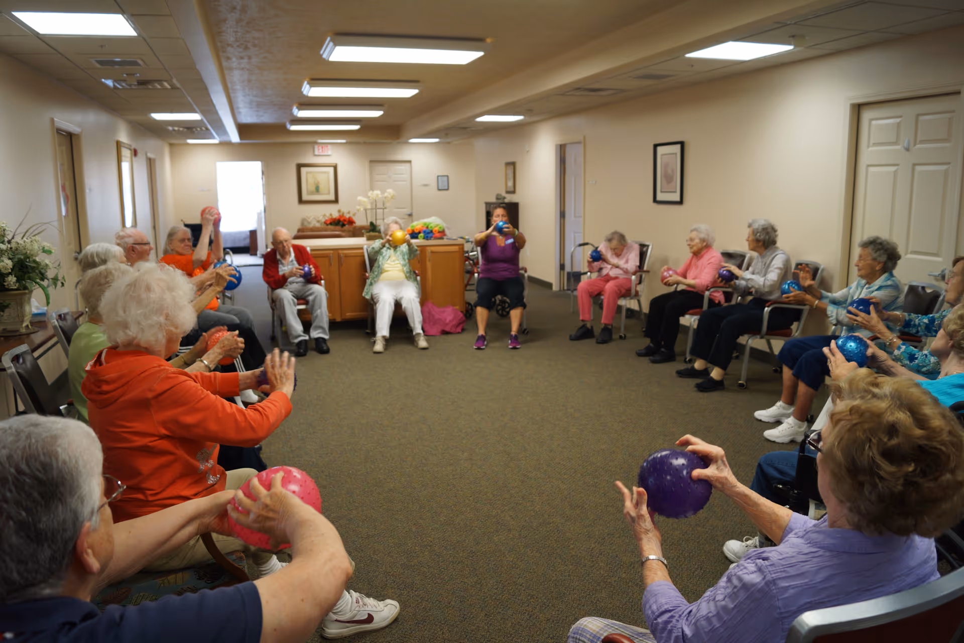 A group of elderly people sitting in a circle in a room, participating in a seated exercise activity using colorful small balls. The room has beige walls, carpeted floor, fluorescent ceiling lights, and framed pictures on the walls.