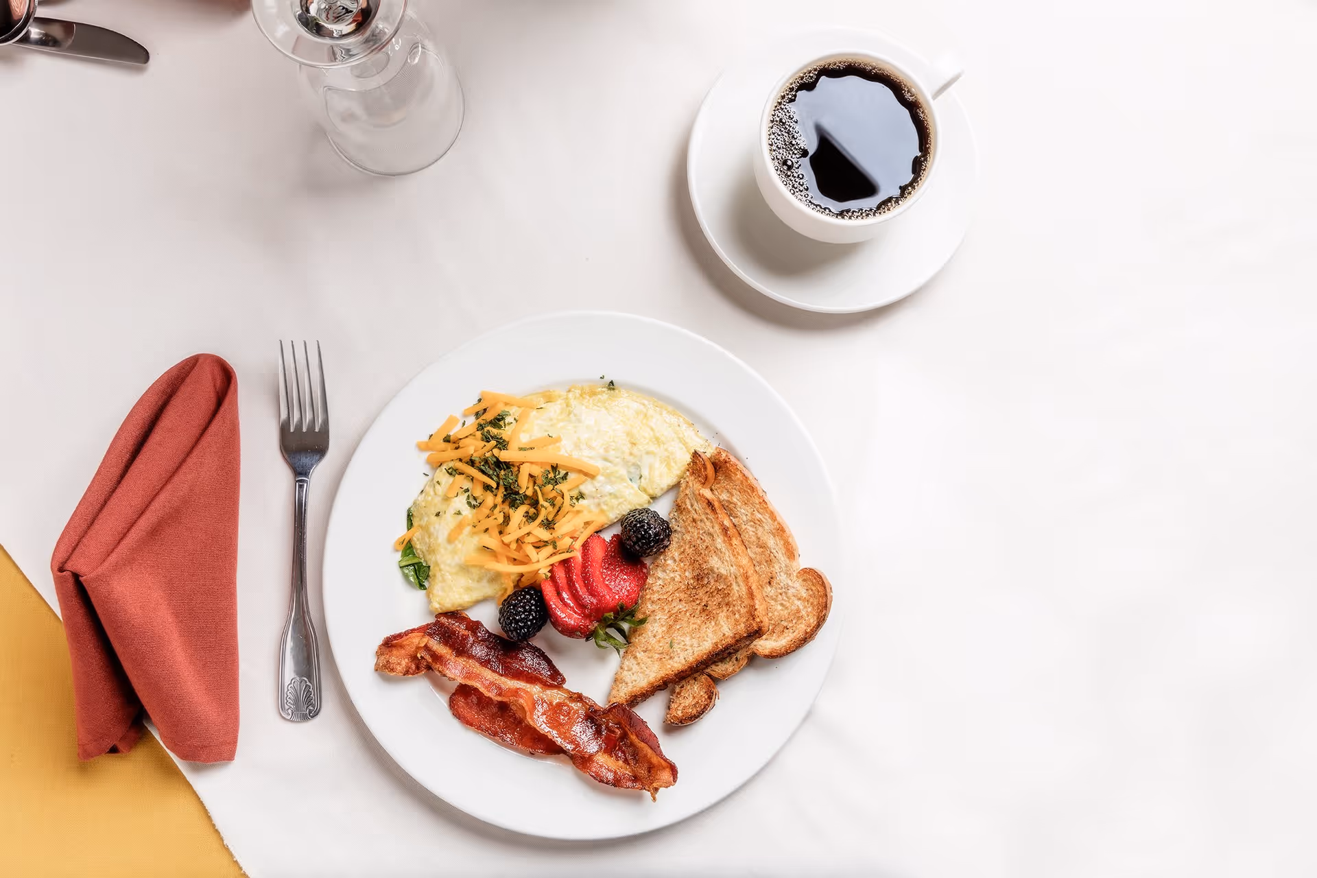 A breakfast plate with an omelet topped with shredded cheese and herbs, two slices of toasted bread, two strips of crispy bacon, and fresh berries including strawberries and blackberries. A cup of black coffee in a white cup and saucer is placed to the right of the plate on a white tablecloth. A fork is placed to the left of the plate next to a folded red napkin.