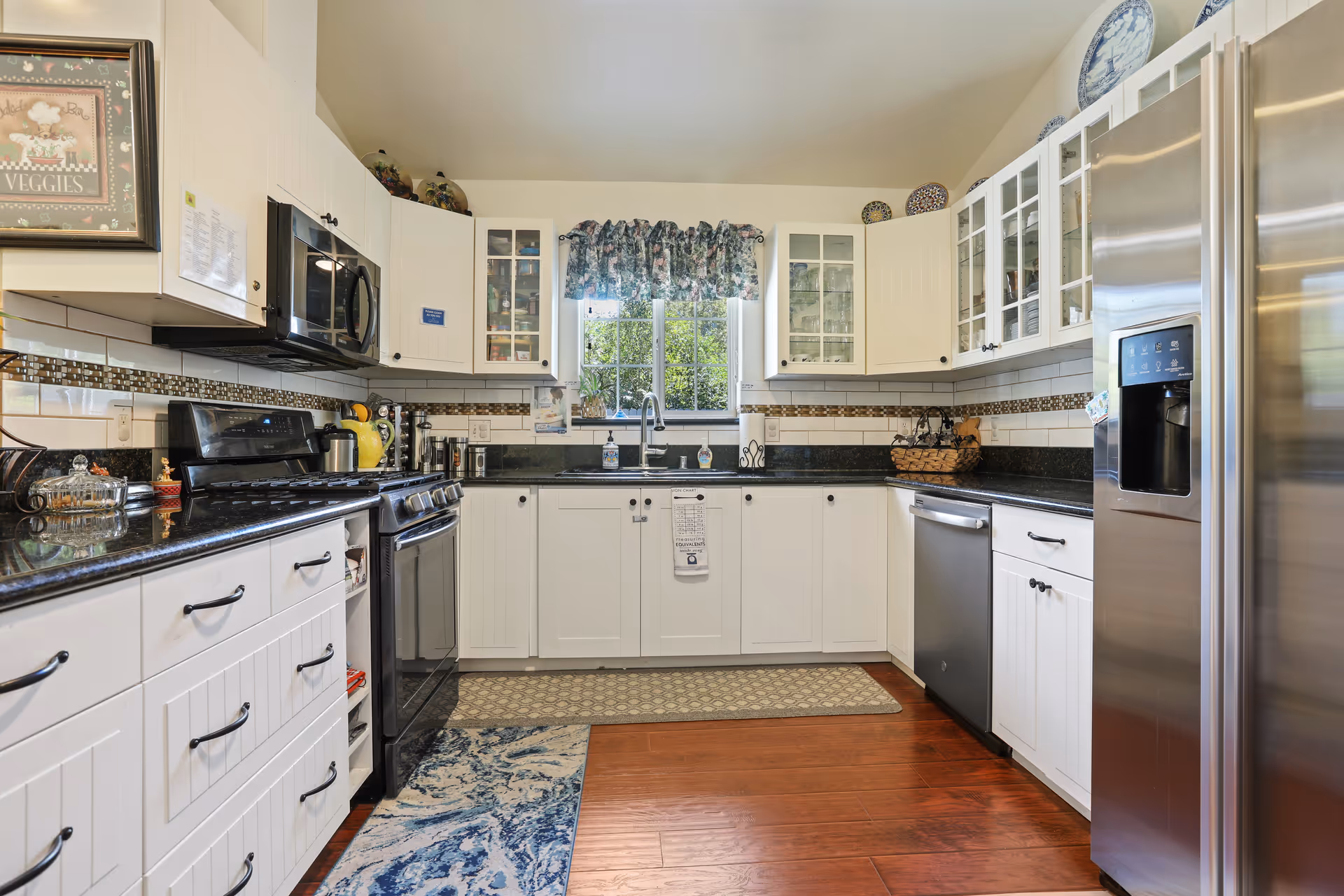 Bright kitchen with white cabinets, black granite countertops, stainless steel appliances including a refrigerator, dishwasher, stove, and microwave. A window with floral curtains is above the sink, and there are decorative plates on top of the cabinets. The floor is wooden with two rugs placed in front of the sink and stove.