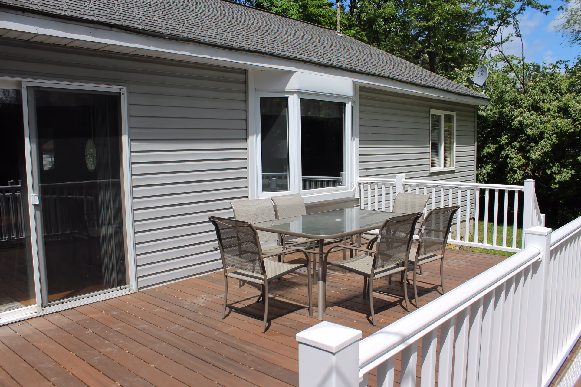 Outdoor wooden deck attached to a house with gray siding. The deck has a glass-top table surrounded by six metal mesh chairs. White railing encloses the deck, and there are trees and greenery in the background under a blue sky with some clouds.