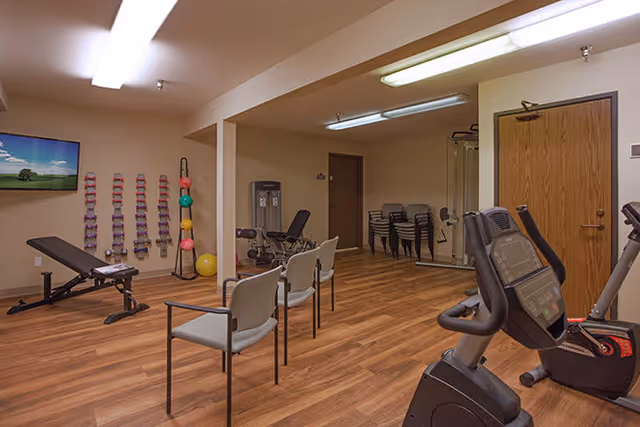 Fitness room with a stationary bike, weight bench, wall-mounted dumbbells and exercise balls, chairs, and wood-look flooring.